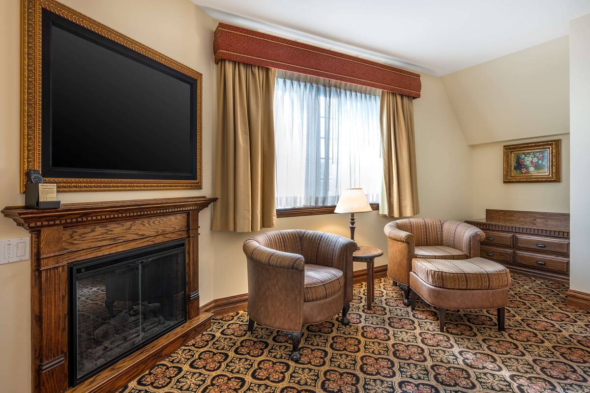 A room with a fireplace, two armchairs, a side table, a lamp, a television, and a painting on the wall.
