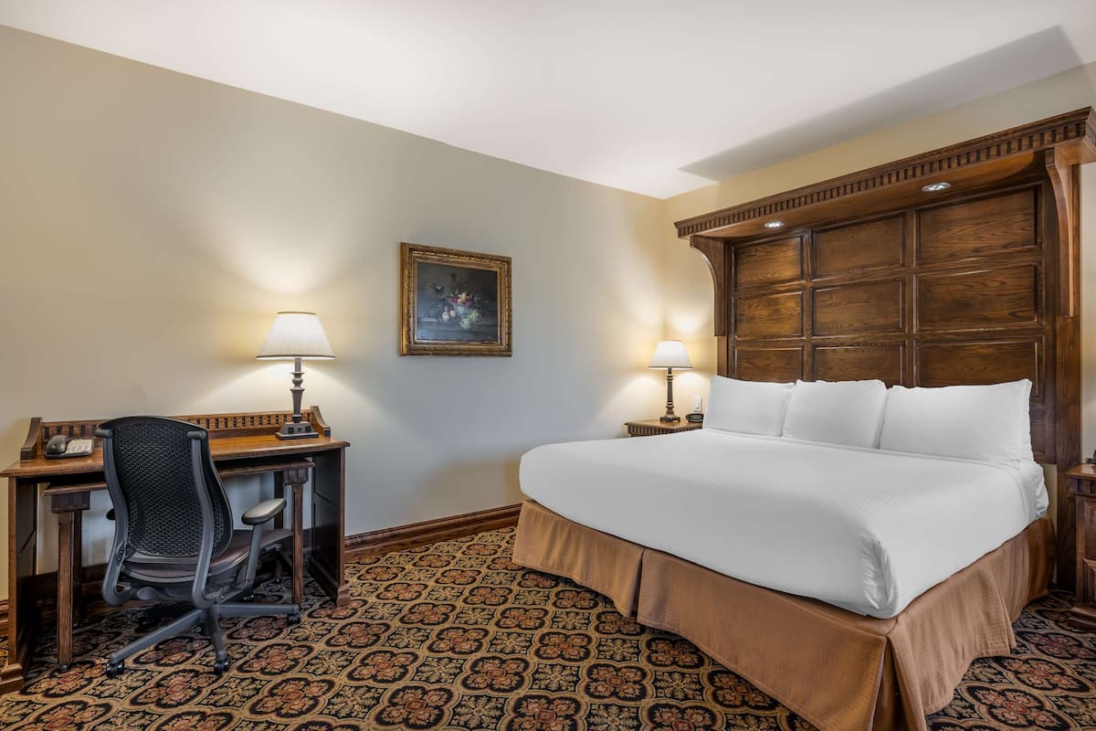 A hotel room with a large bed, a desk with a chair, two bedside lamps, and a patterned carpet.