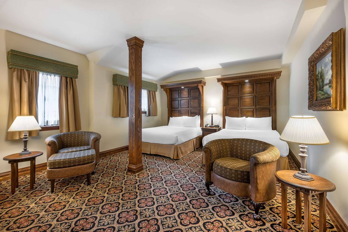 A hotel room with two beds, a large wooden pillar, patterned carpet, and classic furniture.