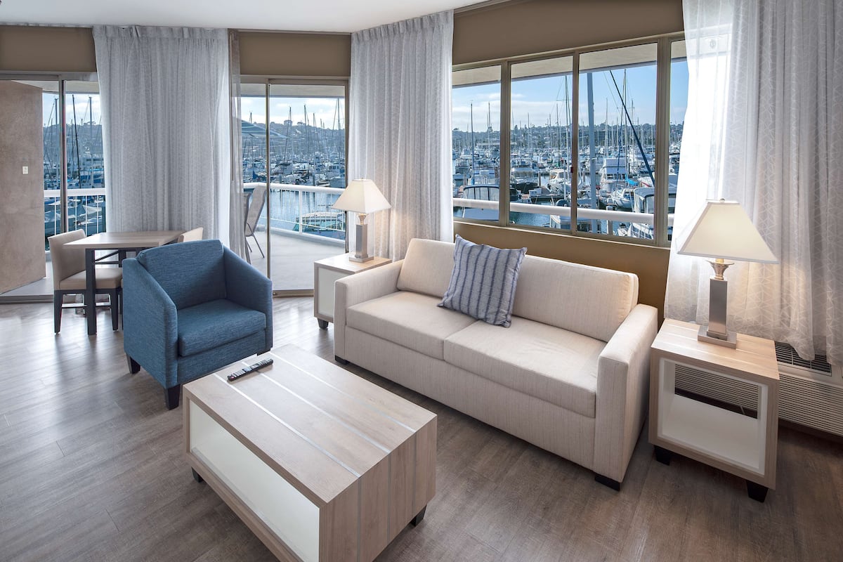 A modern hotel room with a balcony, a sofa, a chair, a coffee table, and two bedside tables with lamps.
