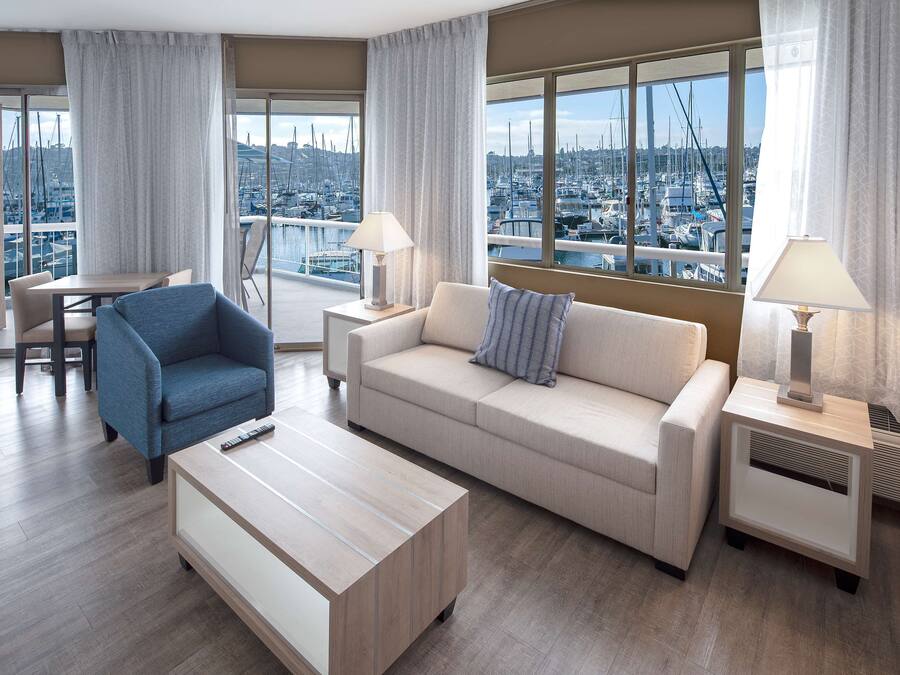 A modern hotel room with a balcony, a sofa, a chair, a coffee table, and two bedside tables with lamps.