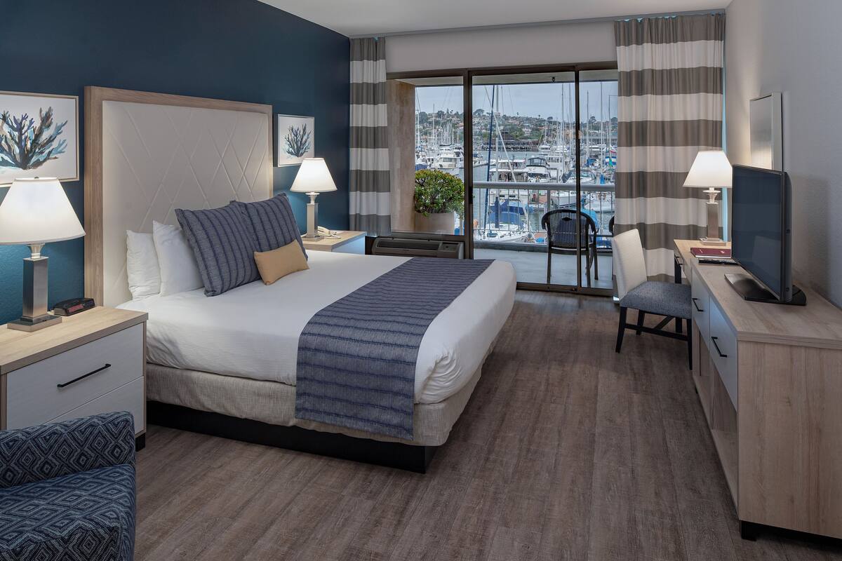 A hotel room with a large bed, two bedside tables with lamps, a desk with a chair, and a view of the marina.