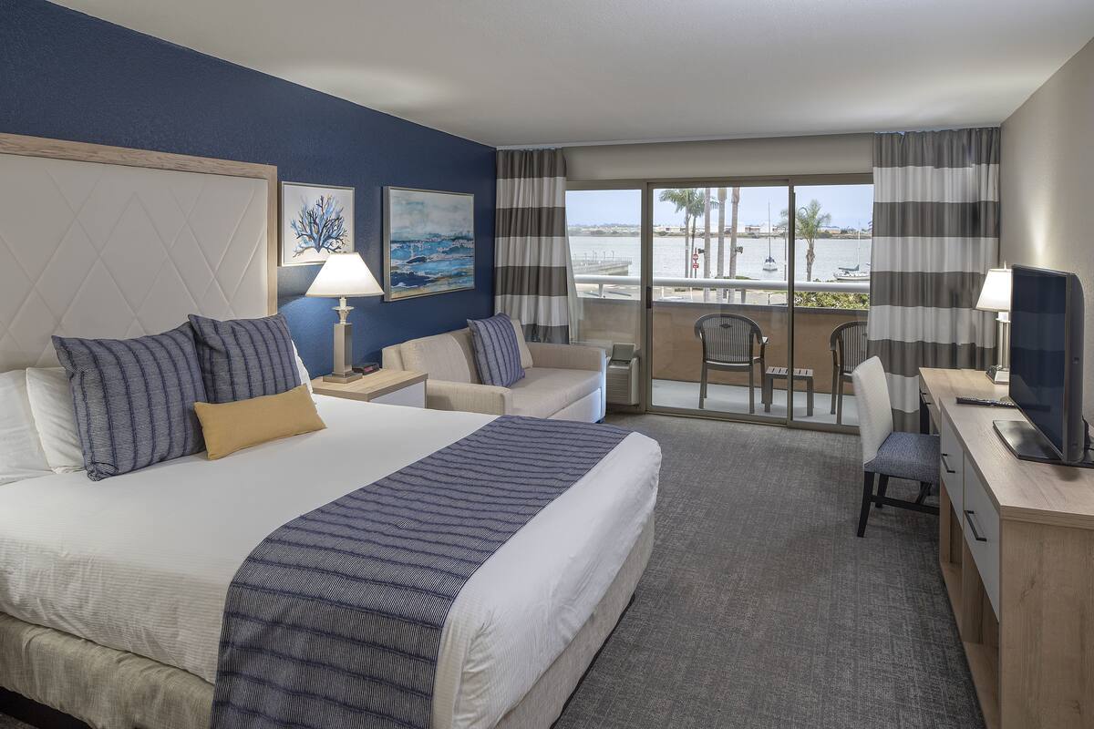 A hotel room with a large bed, a desk, a chair, a balcony with a view of the water and palm trees.