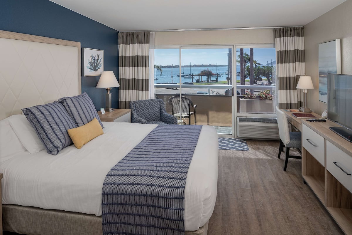 A hotel room with a large bed, a desk with a chair, a TV, and a balcony with a view of the sea and boats.