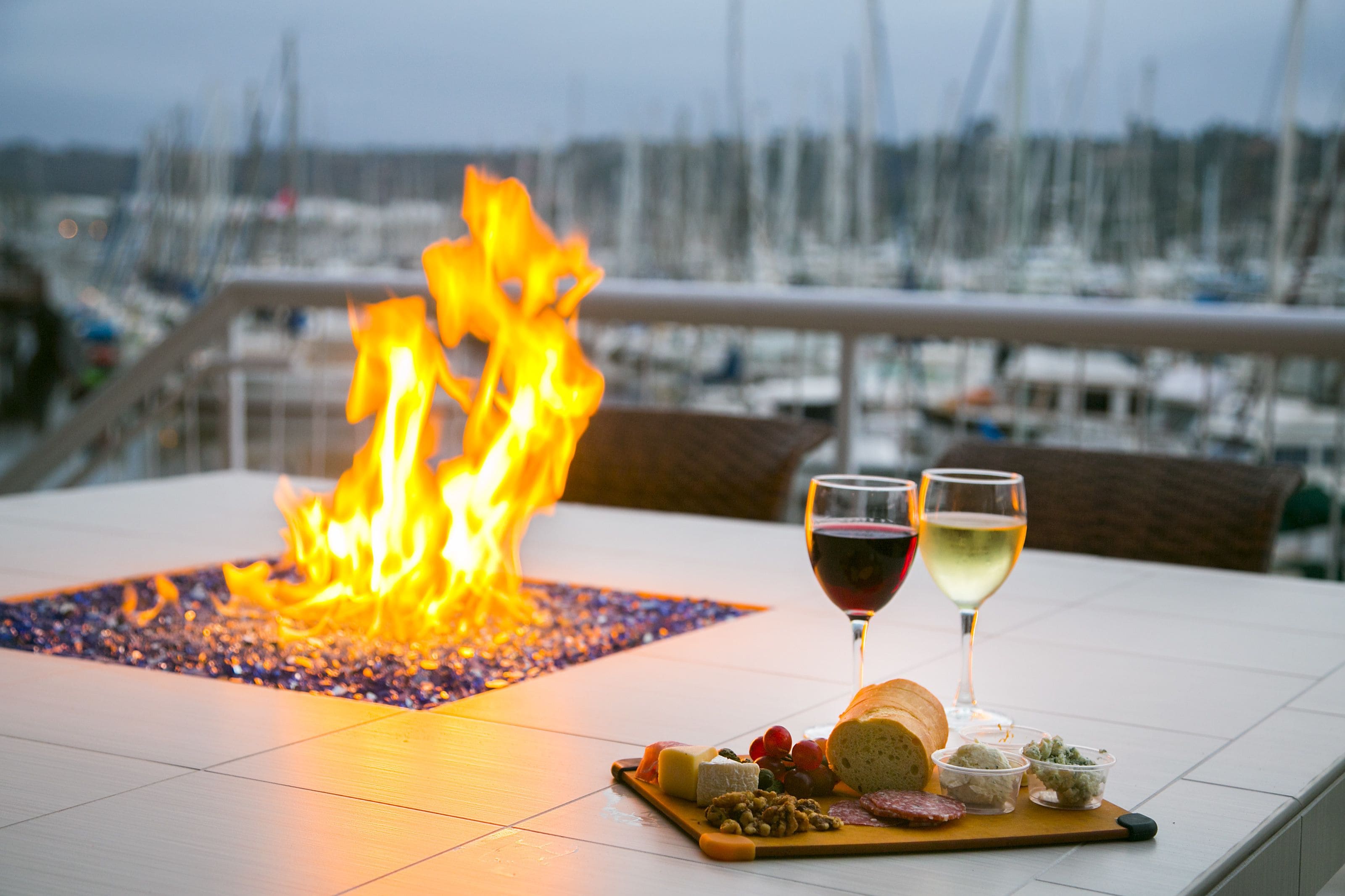 A table with a fire feature, two glasses of wine, and a variety of cheeses and bread.