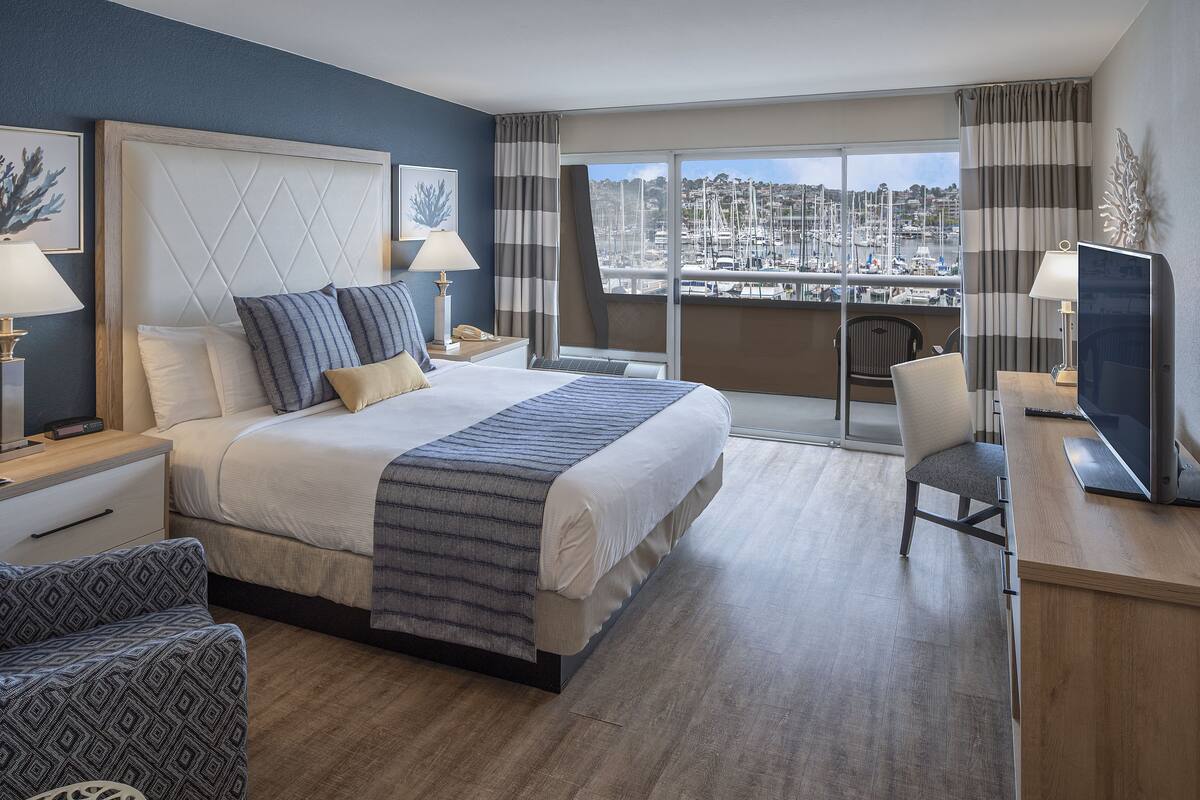 A hotel room with a large bed, a desk with a computer, a chair, and a view of a marina.