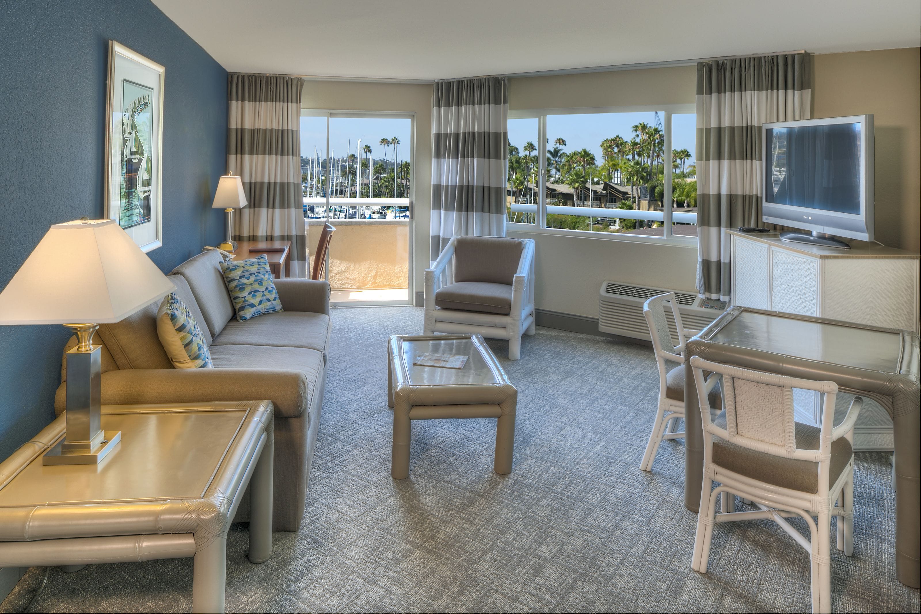 A hotel room with a sofa, armchair, coffee table, TV, and a view of palm trees and a marina.