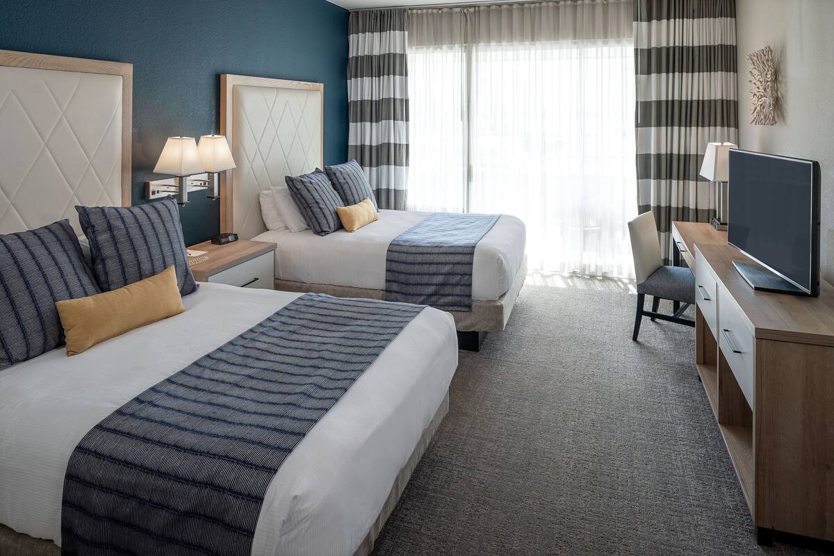 A hotel room with two beds, a desk, a TV, and a window with curtains.