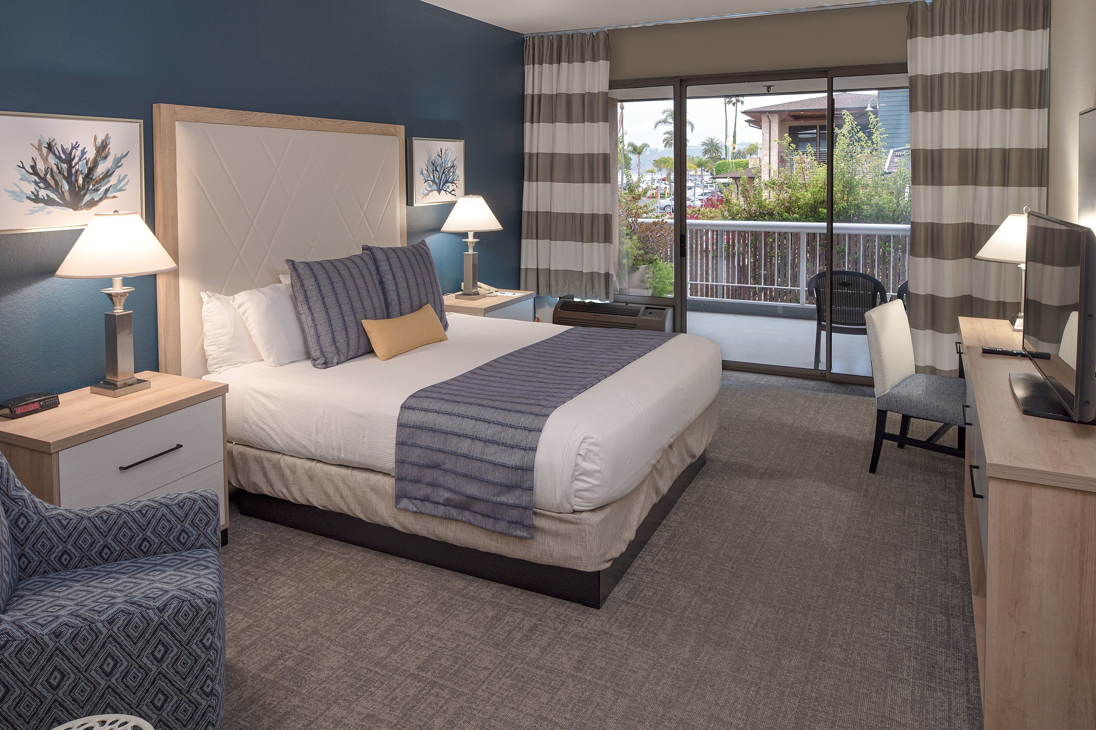A hotel room with a large bed, two bedside tables with lamps, a desk with a chair, and a view of the outside balcony.