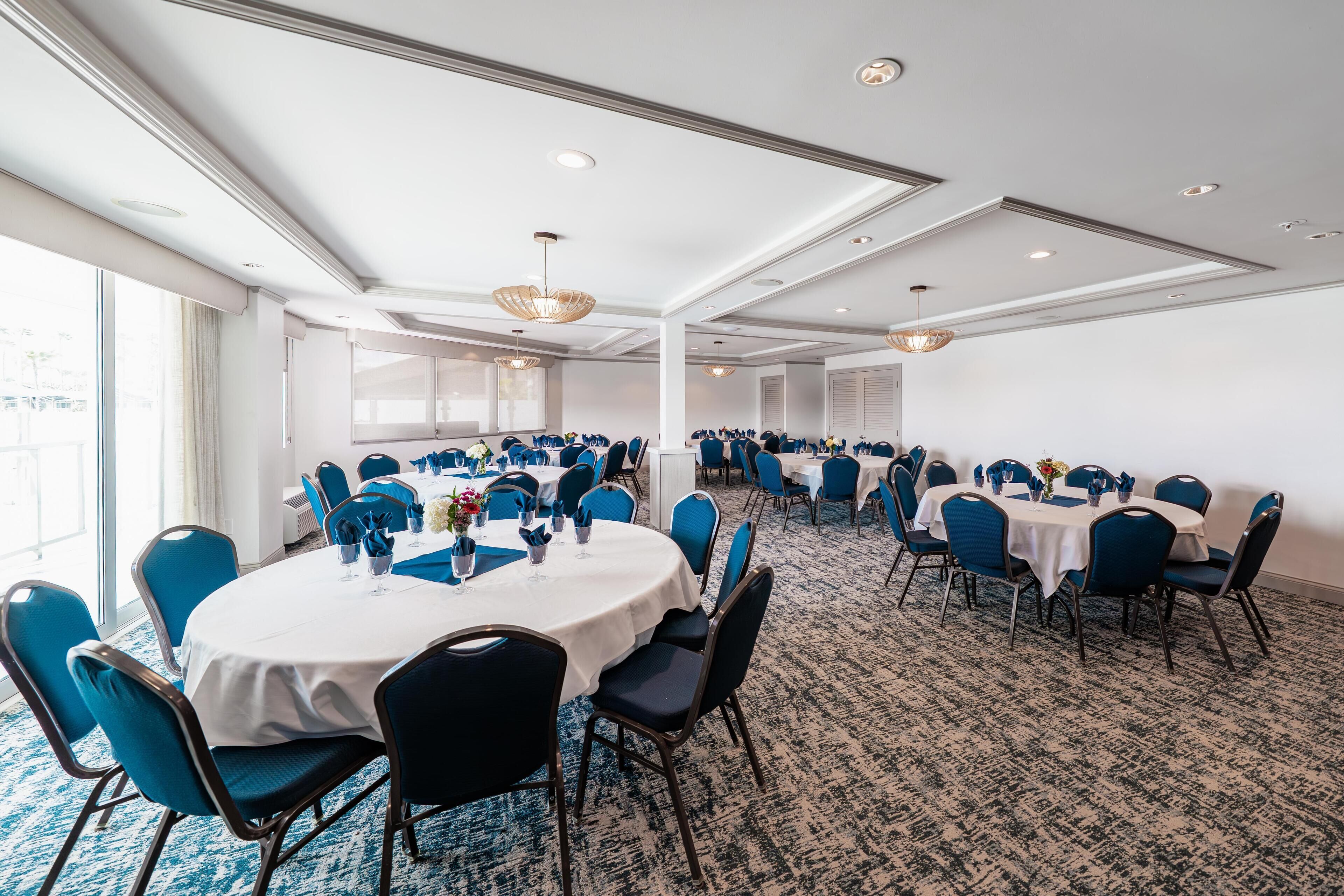 Une salle de conférence avec des tables rondes et des chaises bleues, un sol recouvert de moquette et de grandes fenêtres.