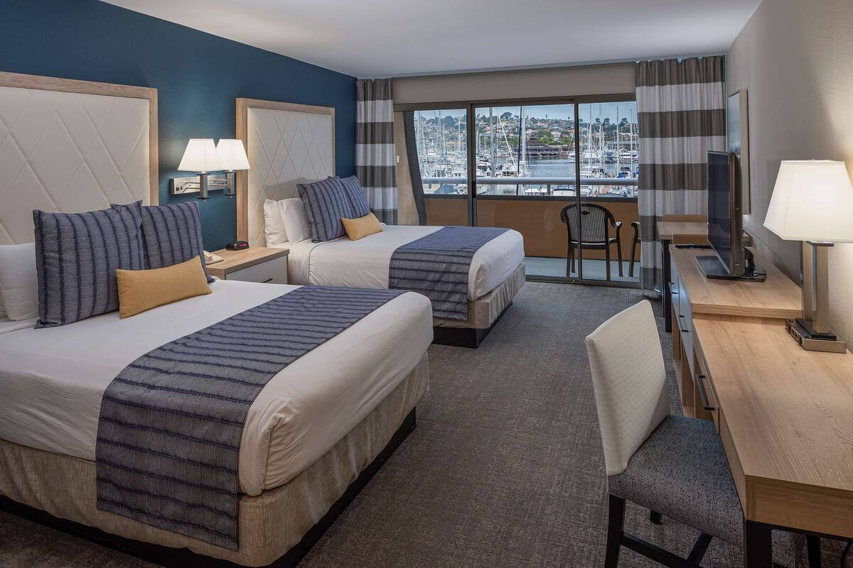 A hotel room with two beds, a desk, a chair, and a large window with a view of a marina.