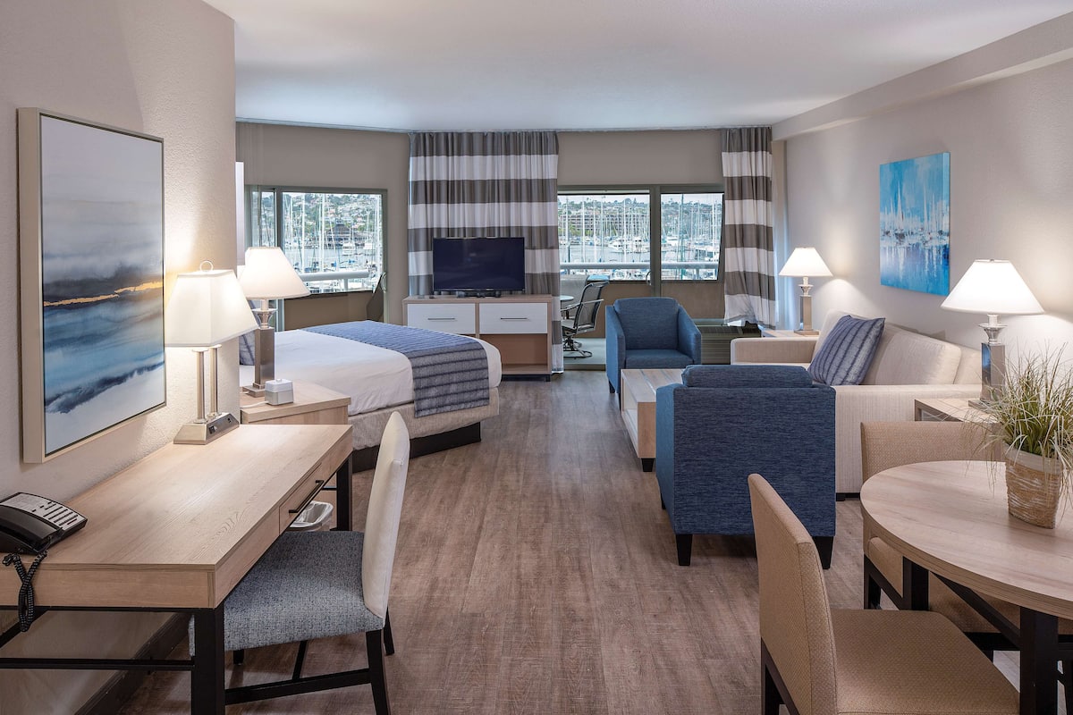 A hotel room with a bed, desk, chair, sofa, TV, and large windows with a city view.