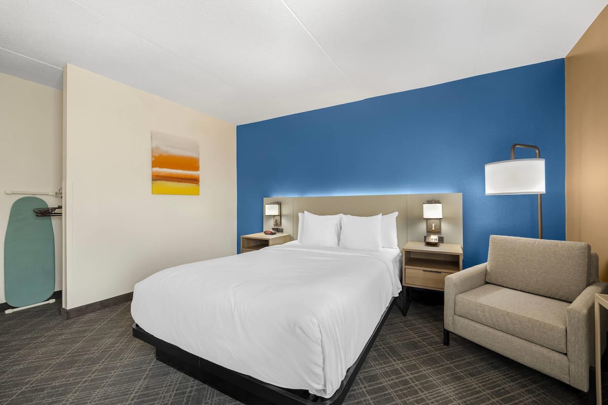 A hotel room with a large bed, a chair, a nightstand with a lamp, and a wall-mounted artwork.