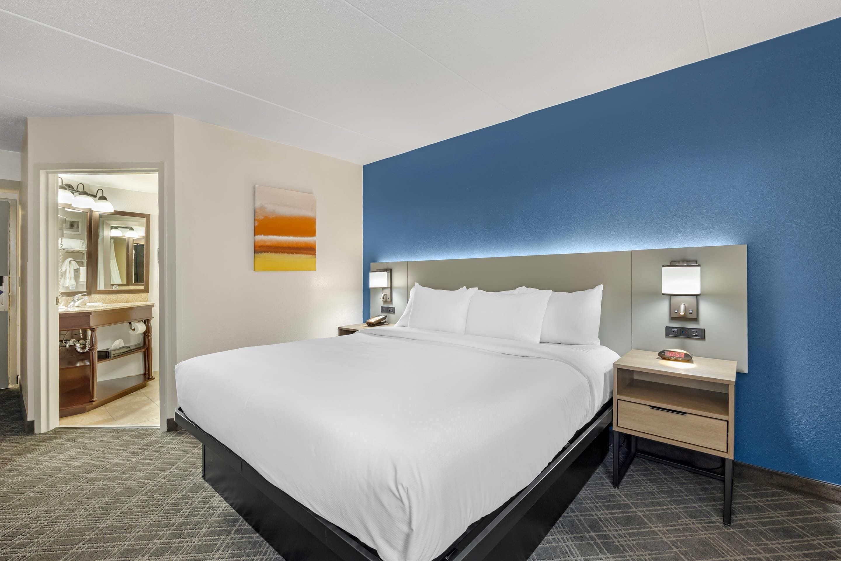Ein Hotelzimmer mit einem großen Bett, einem Nachttisch mit Lampe, einer an der Wand befestigten Ablage und einem durch eine offene Tür einsehbaren Badezimmer.