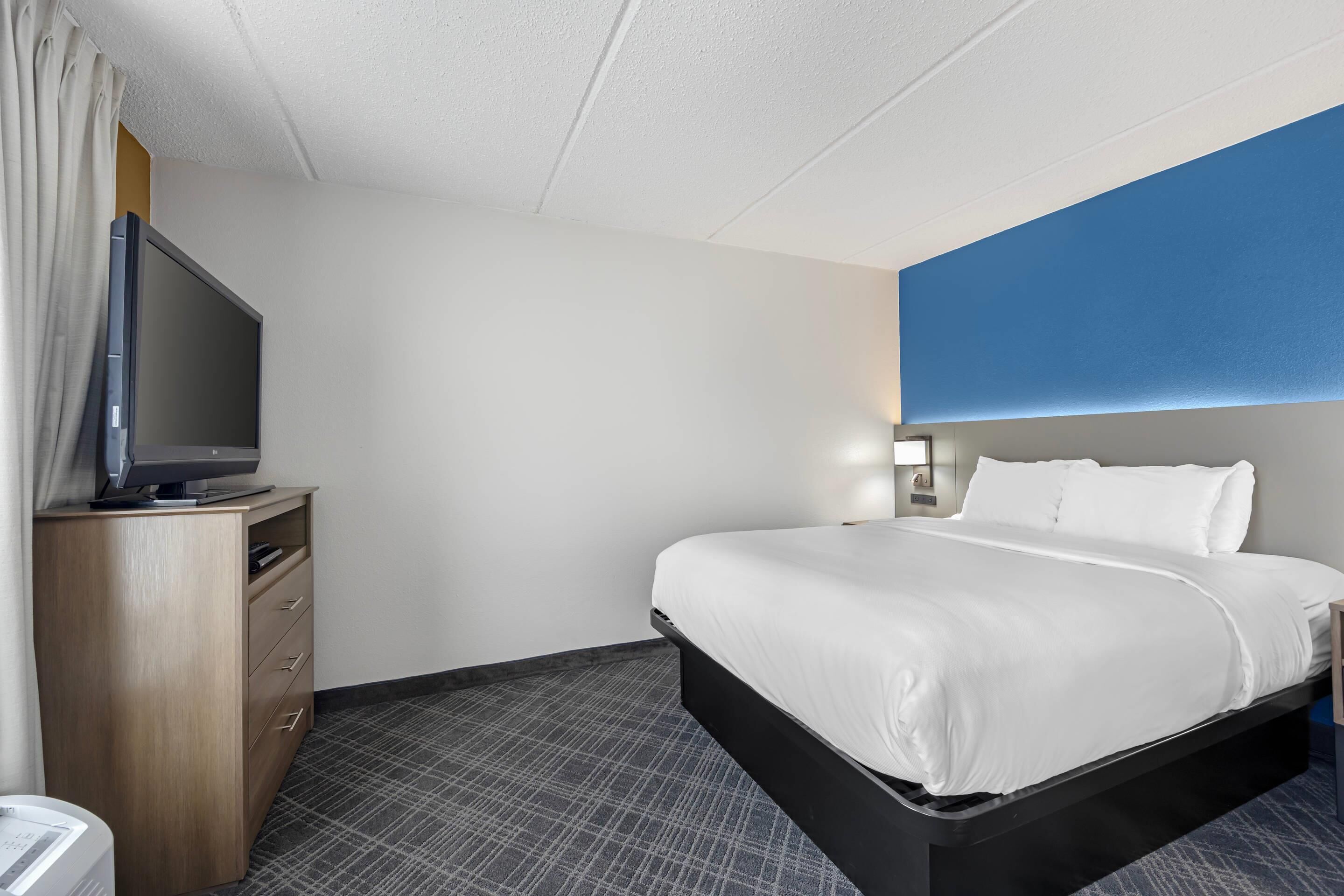 A hotel room with a bed, a television on a wooden dresser, and a blue accent wall.