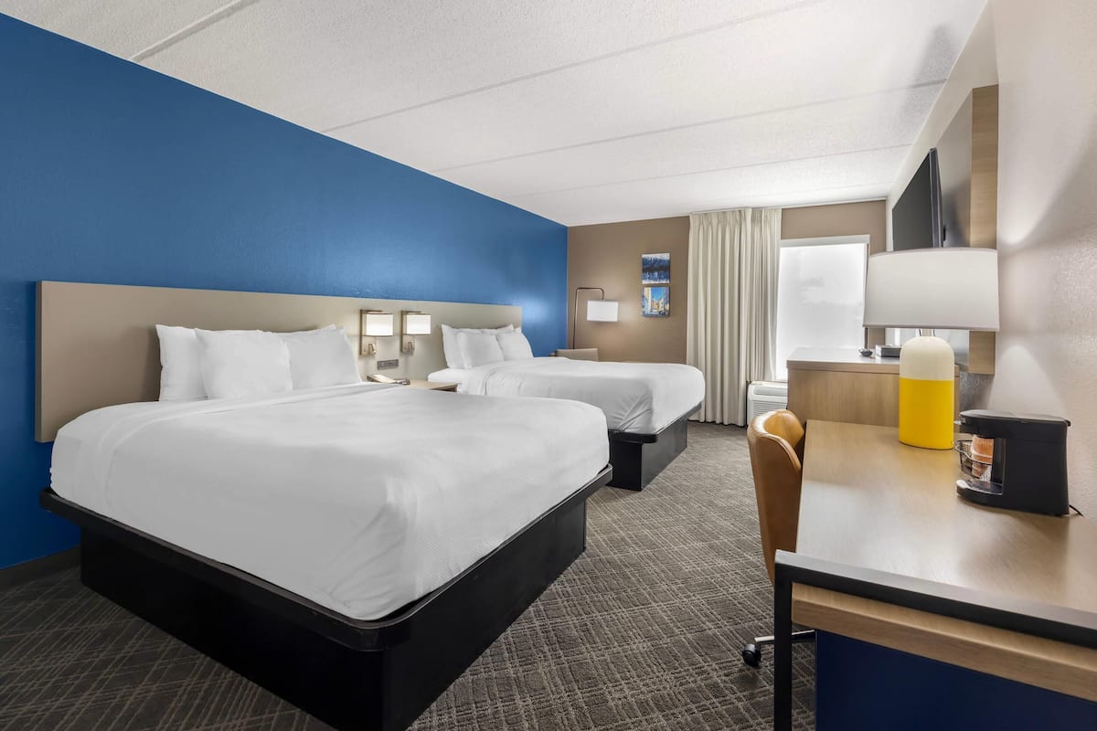 A hotel room with a large bed, two smaller beds, a desk with a lamp, a chair, and a TV.