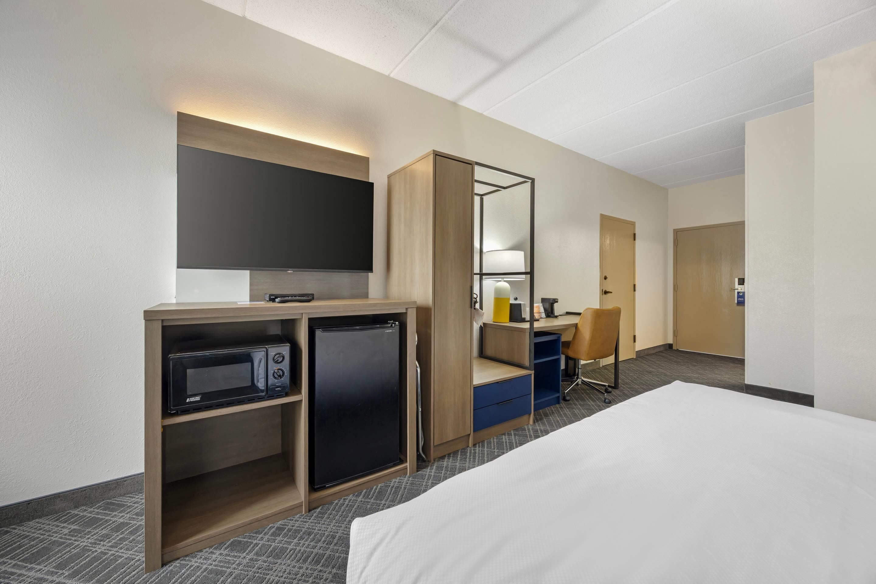 A hotel room with a bed, a desk with a chair, a microwave, a refrigerator, a TV, and a large mirror.