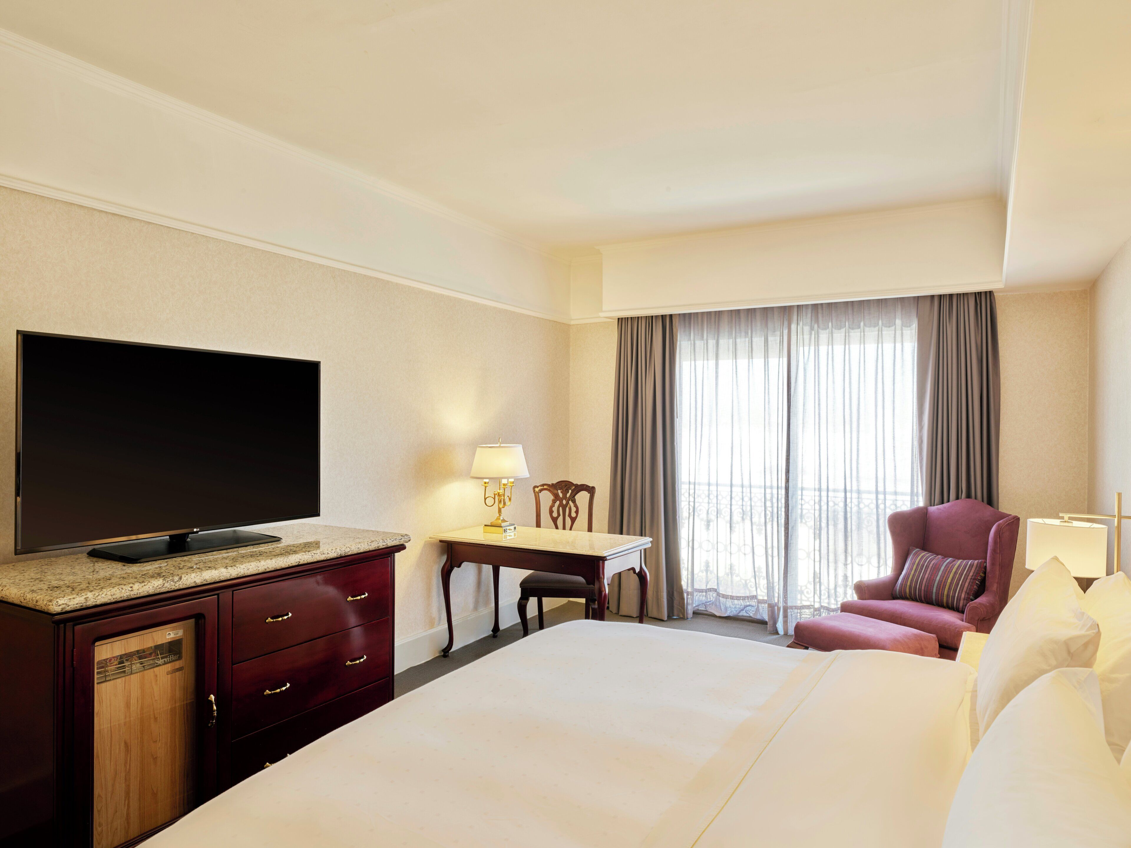 A hotel room with a bed, a television, a desk with a lamp, a chair, and a window with curtains.