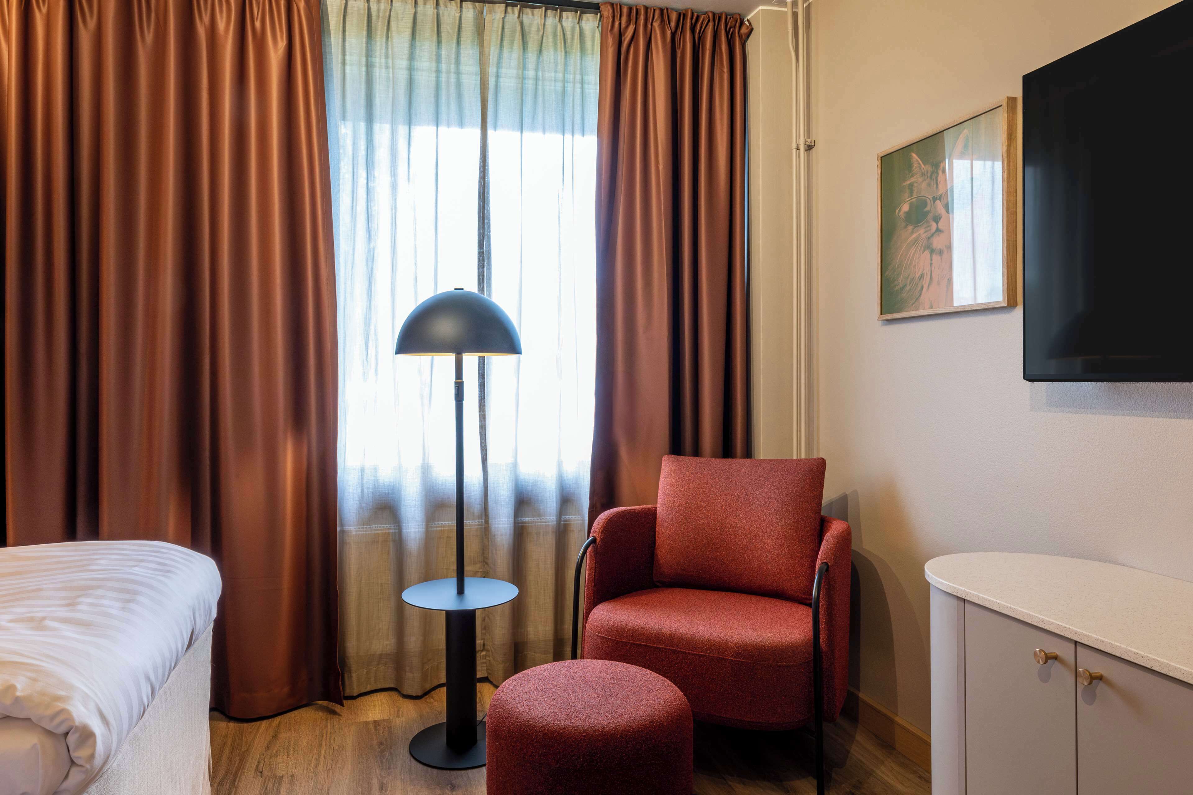 Et hotelværelse med en seng, en rød lænestol, en lille rund skammel, en gulvlampe, et fjernsyn på væggen og et vindue med gardiner.