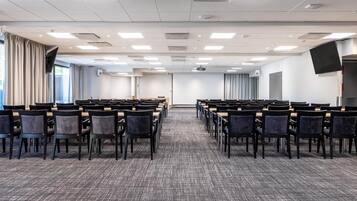 Un salón de conferencias con hileras de sillas, suelo alfombrado, luces de techo y una pantalla de proyector.