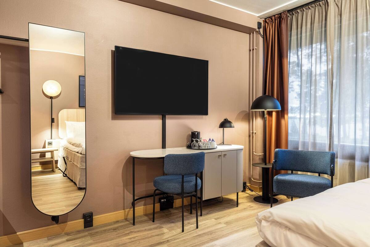 Ein Hotelzimmer mit Bett, Schreibtisch mit Stuhl, Fernseher, Lampe und einem Spiegel, der den Raum widerspiegelt.