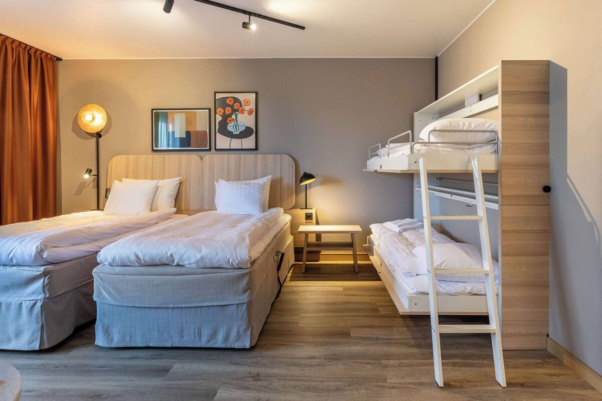 Ein modernes Hotelzimmer mit zwei Betten, einem Etagenbett, einem Nachttisch, einer Lampe und einem Wandregal.