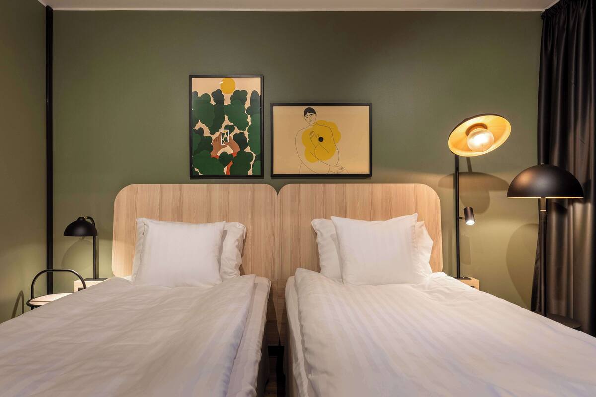 Ein Hotelzimmer mit zwei Betten, einem Holzkopfteil, einem Nachttisch, einer Lampe und zwei gerahmten Kunstwerken an der Wand.