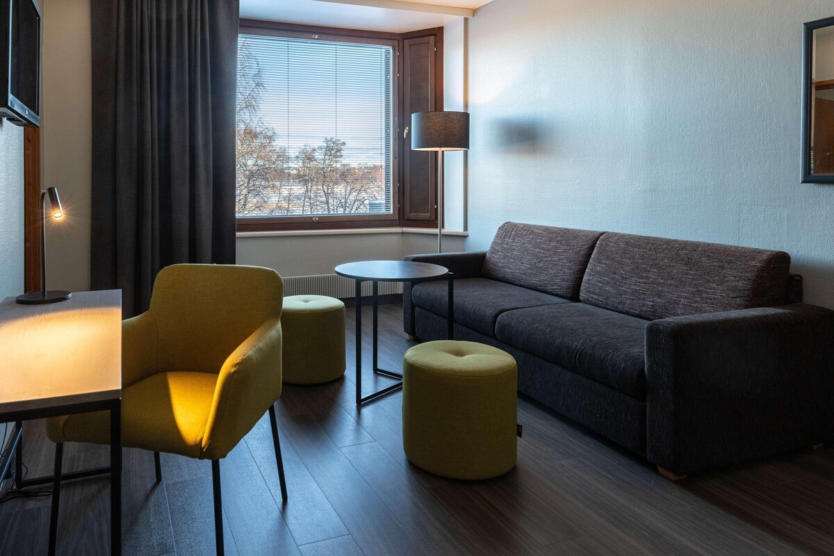 A modern living room with a sofa, two ottomans, a small table, a lamp, and a window with blinds.