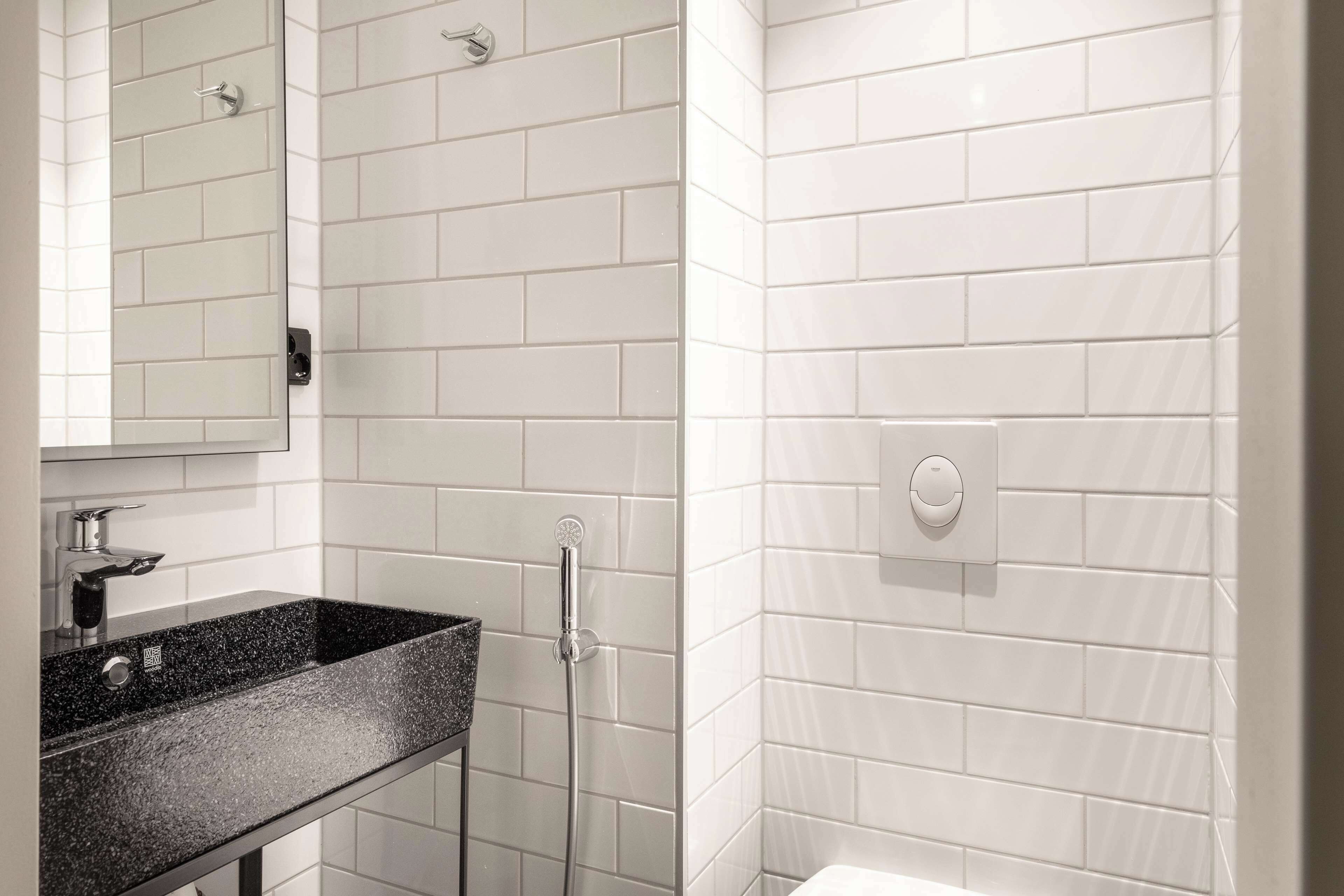 Ein Badezimmer mit weißen Fliesen im U-Bahn-Stil, einem schwarzen Granitwaschbecken, einem Spiegel, einer Handbrause und einer Toilette.