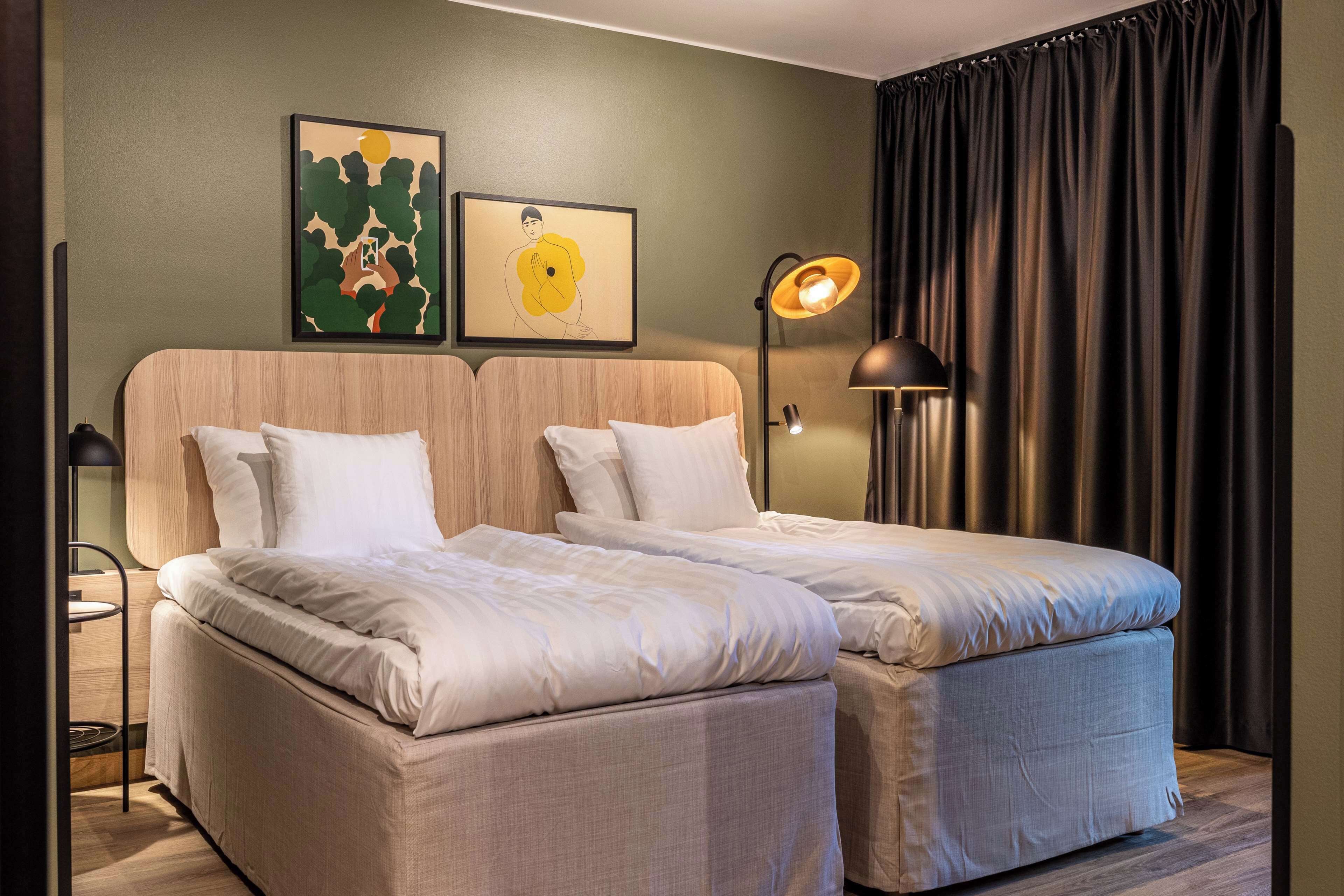 A hotel room with two beds, a headboard, a bedside table, a lamp, and artwork on the wall.
