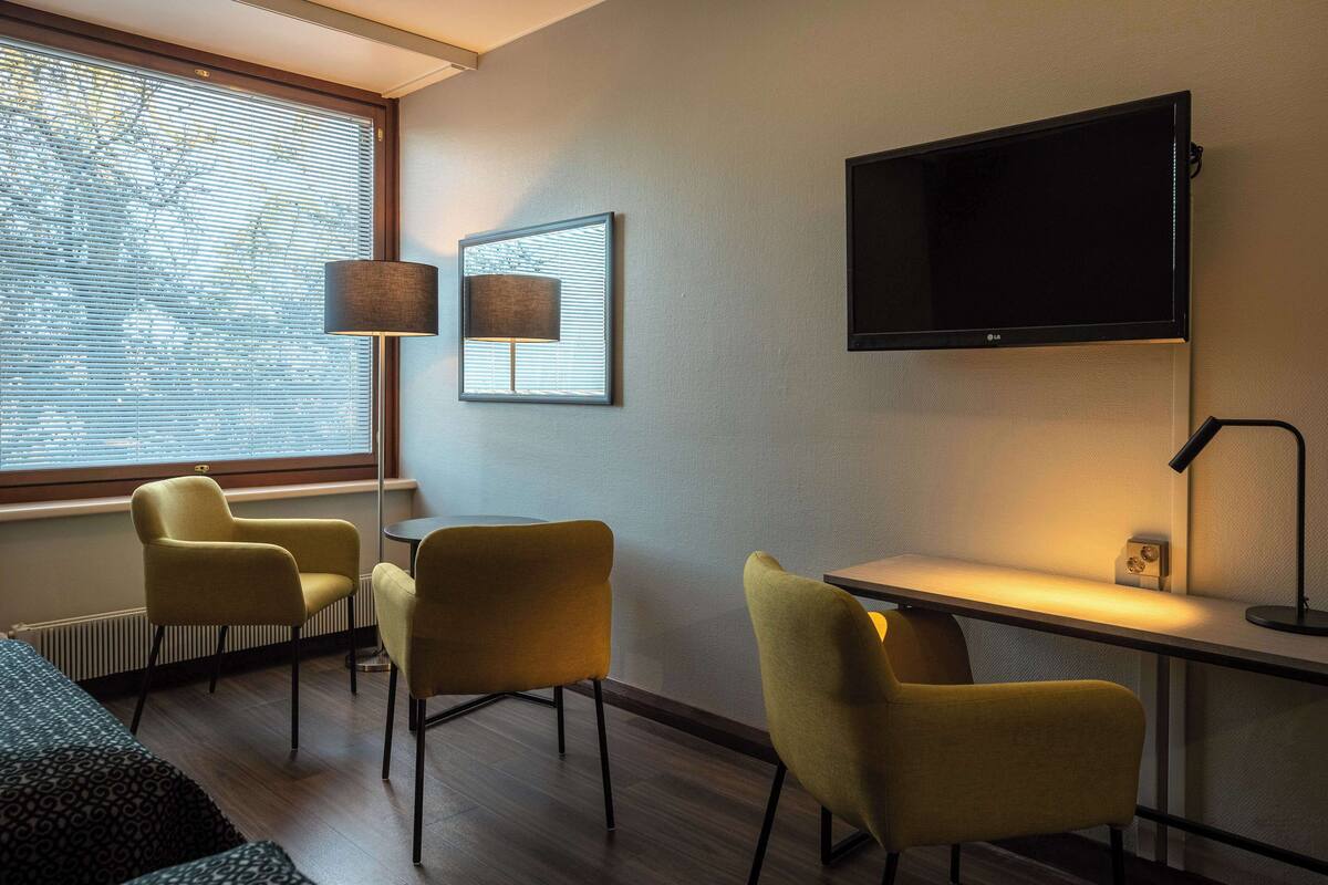 Ein modernes Hotelzimmer mit großem Fenster, zwei gelben Stühlen, einem Schreibtisch mit Lampe und einem am Wand montierten Flachbildfernseher.