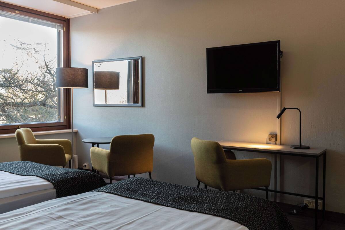 Ein Hotelzimmer mit einem Bett, zwei Stühlen, einem Schreibtisch, einem Fernseher und einem Fenster mit Blick auf Bäume.