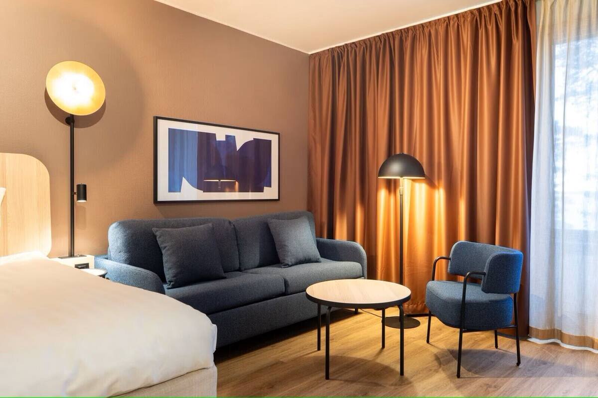 Ein Hotelzimmer mit Bett, Sofa, Sessel, kleinem Tisch, Stehlampe, Wandbild und Vorhängen.