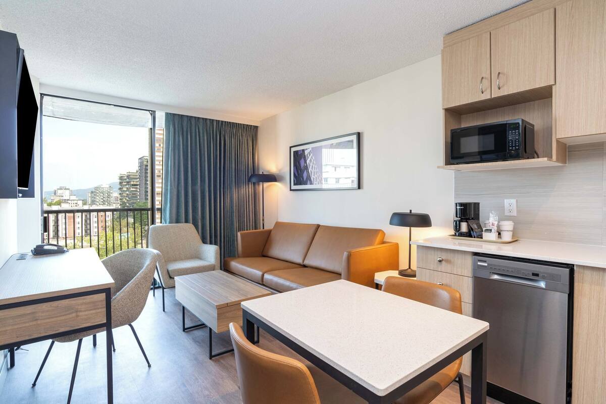 Una habitación de hotel moderna con balcón, sofá, mesa de comedor y una zona de cocina equipada con microondas y lavavajillas.