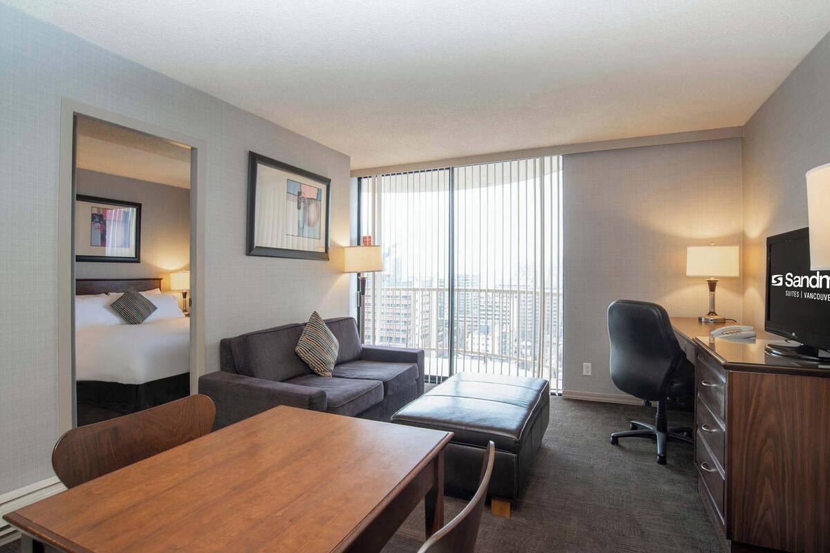 Habitación de hotel con cama, sofá, escritorio, televisión y ventana con persianas.