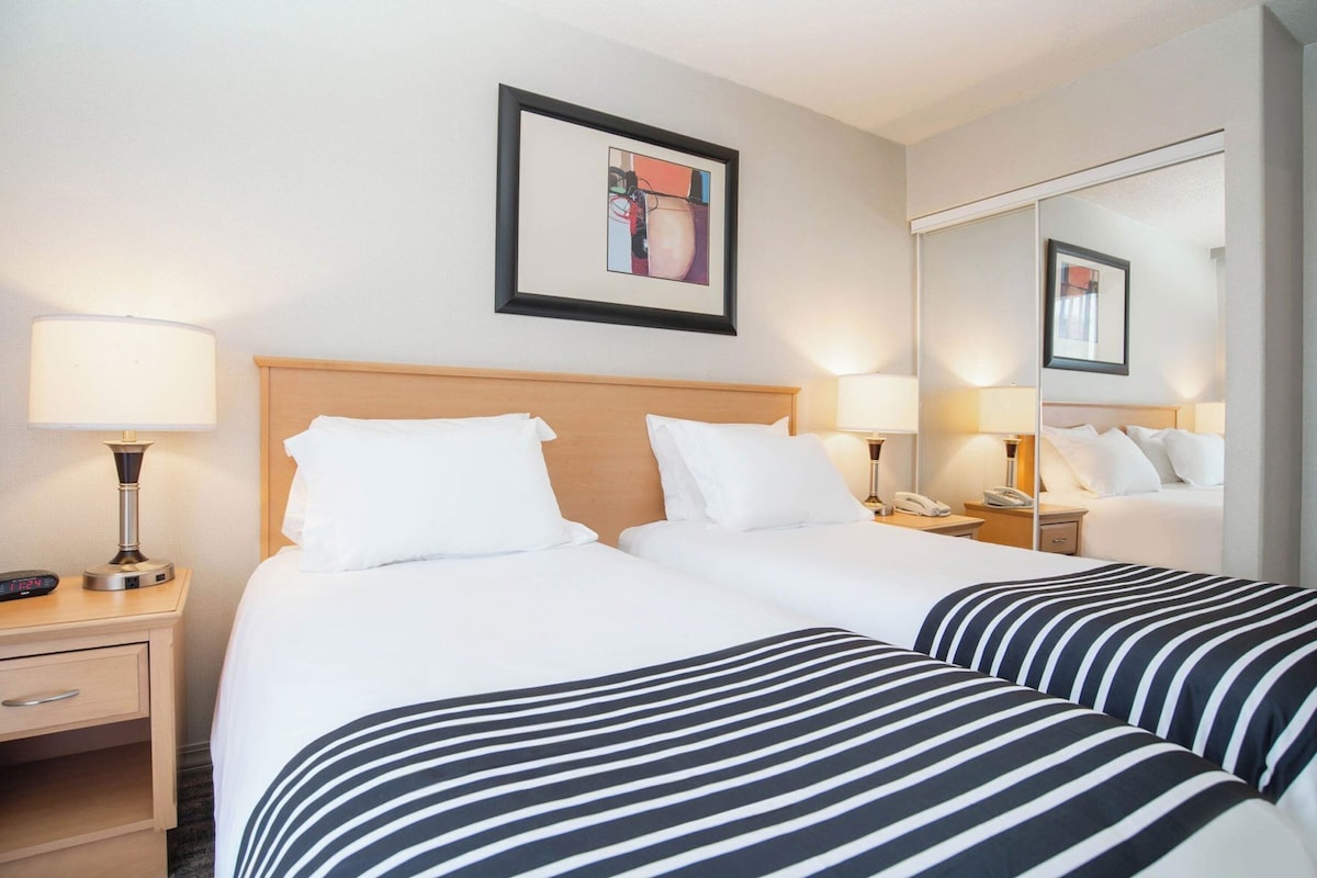 Une chambre d’hôtel avec deux lits, une tête de lit en bois, des lampes de chevet, un téléphone sur la table de chevet, un tableau encadré au mur et un grand miroir.