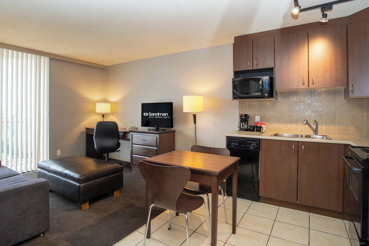 Habitación de hotel compacta con kitchenette, zona de comedor con mesa y sillas, sofá, televisor y lámpara.