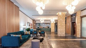 Een moderne lobby met een blauwe bank, een rond poefje en een salontafel. Er staan twee stoelen en een klein tafeltje met een lamp. De vloer is betegeld, en er zijn stenen pilaren.