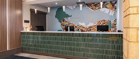Una mesa de recepción con un mural de paisaje urbano, un mostrador de azulejos verdes y un letrero que dice "Sandman Suites".
