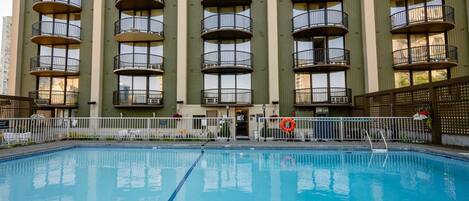 Una piscina con una linea d'acqua blu trasparente, circondata da un recinto bianco. Sullo sfondo, un edificio a più piani con balconi e finestre.