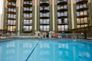 A swimming pool with a clear blue waterline, surrounded by a white fence. In the background, there is a multi-story building with balconies and windows.