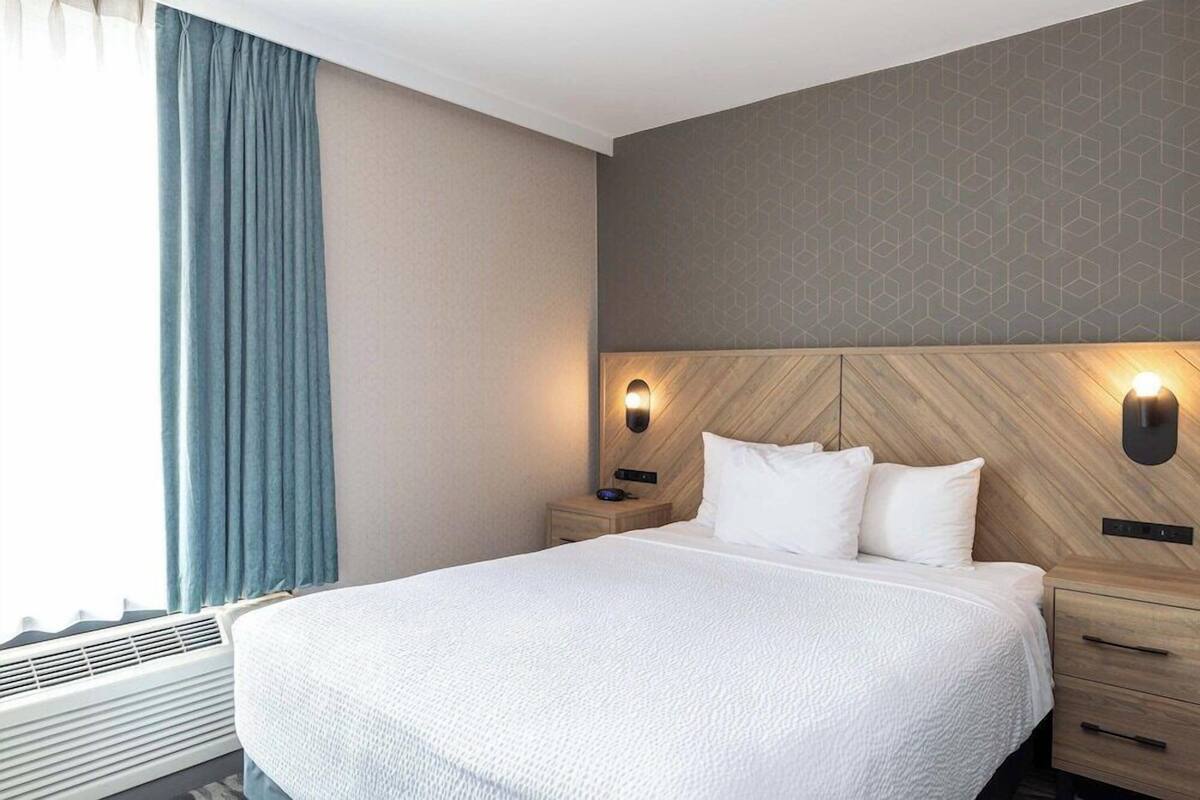 Una habitación de hotel con una cama grande, mesitas de noche, una lámpara de pared y un ventanal con cortinas.