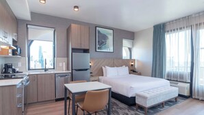 Een hotelkamer met een bed, een bureau, een stoel, een kitchenette en een groot raam met uitzicht op de stad.
