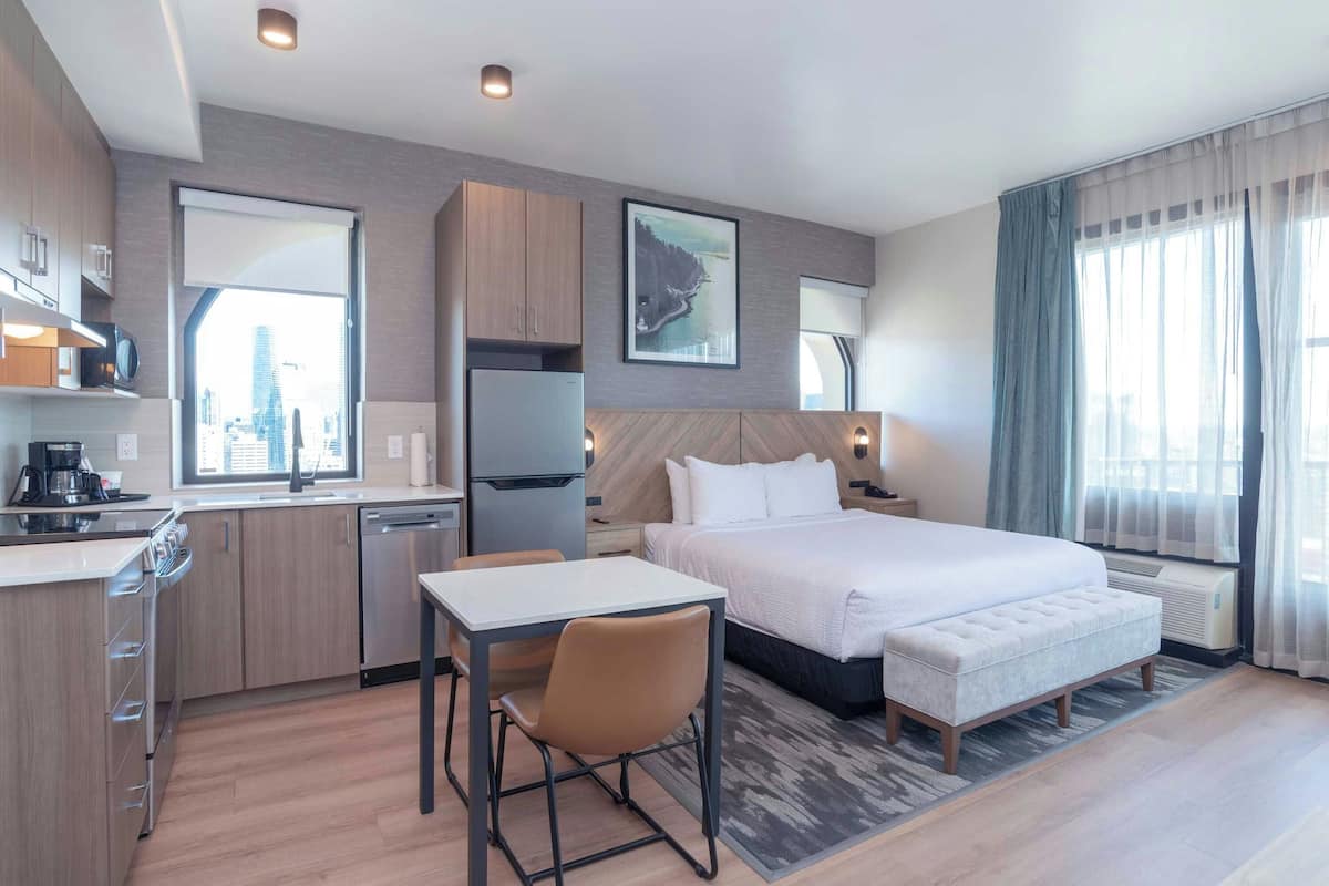 Una habitación de hotel con cama, escritorio, silla, kitchenette y un gran ventanal con vistas a la ciudad.