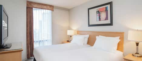 Quarto de hotel com cama grande, televisão em escrivaninha, abajur e janela com cortinas.