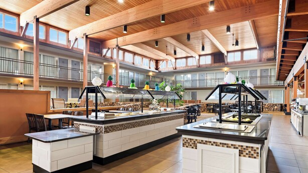 A spacious indoor area with a high ceiling and wooden beams, featuring multiple dining tables and a central counter with a display of colorful items.
