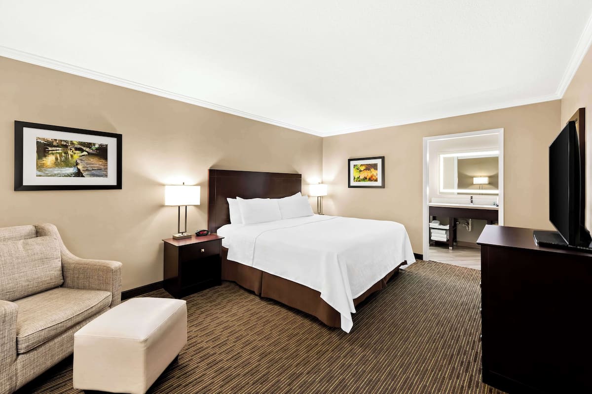 A hotel room with a bed, a chair, a desk, a TV, and a bathroom visible through an open door.