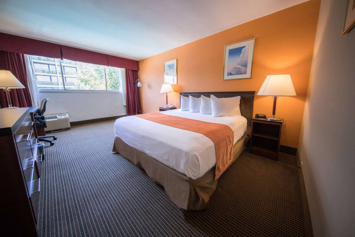 A hotel room with a bed, two bedside lamps, a desk with a chair, and a window with curtains.