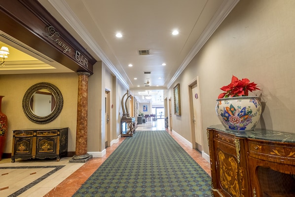 A hotel corridor with a patterned carpet, a lounge bar sign, a decorative mirror, a vase with flowers, and a wooden cabinet.