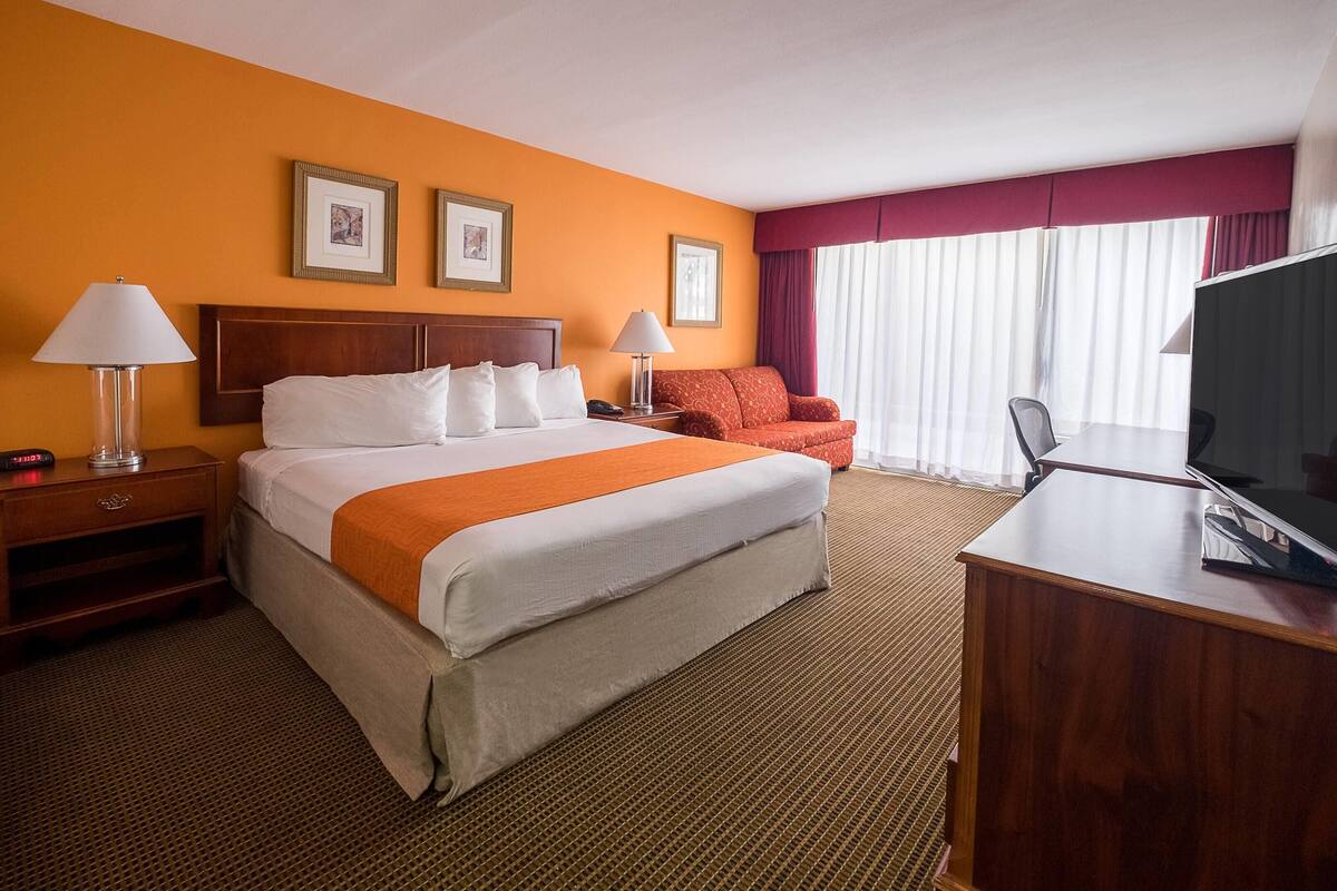 A hotel room with a bed, two bedside tables, a TV, a red armchair, and a window with curtains.